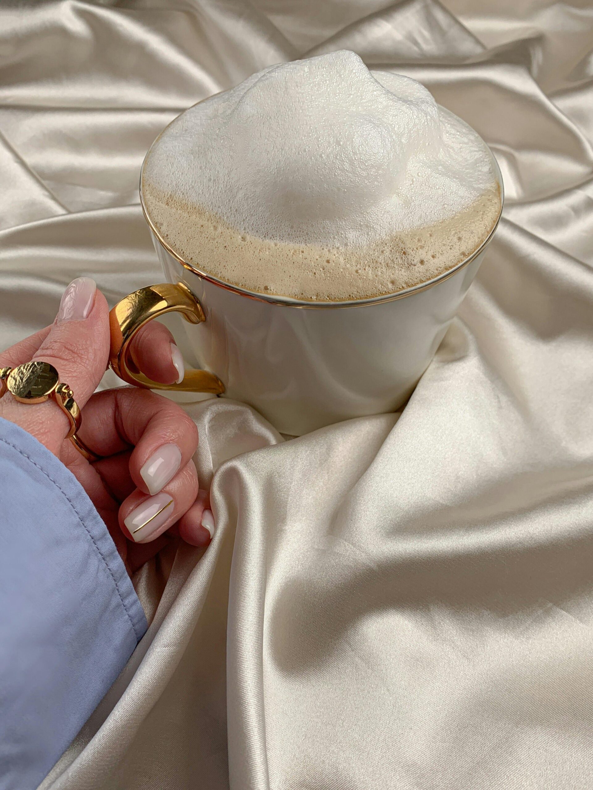 Close-up of a cappuccino in an elegant gold-trimmed cup, held by a well-manicured hand on satin.